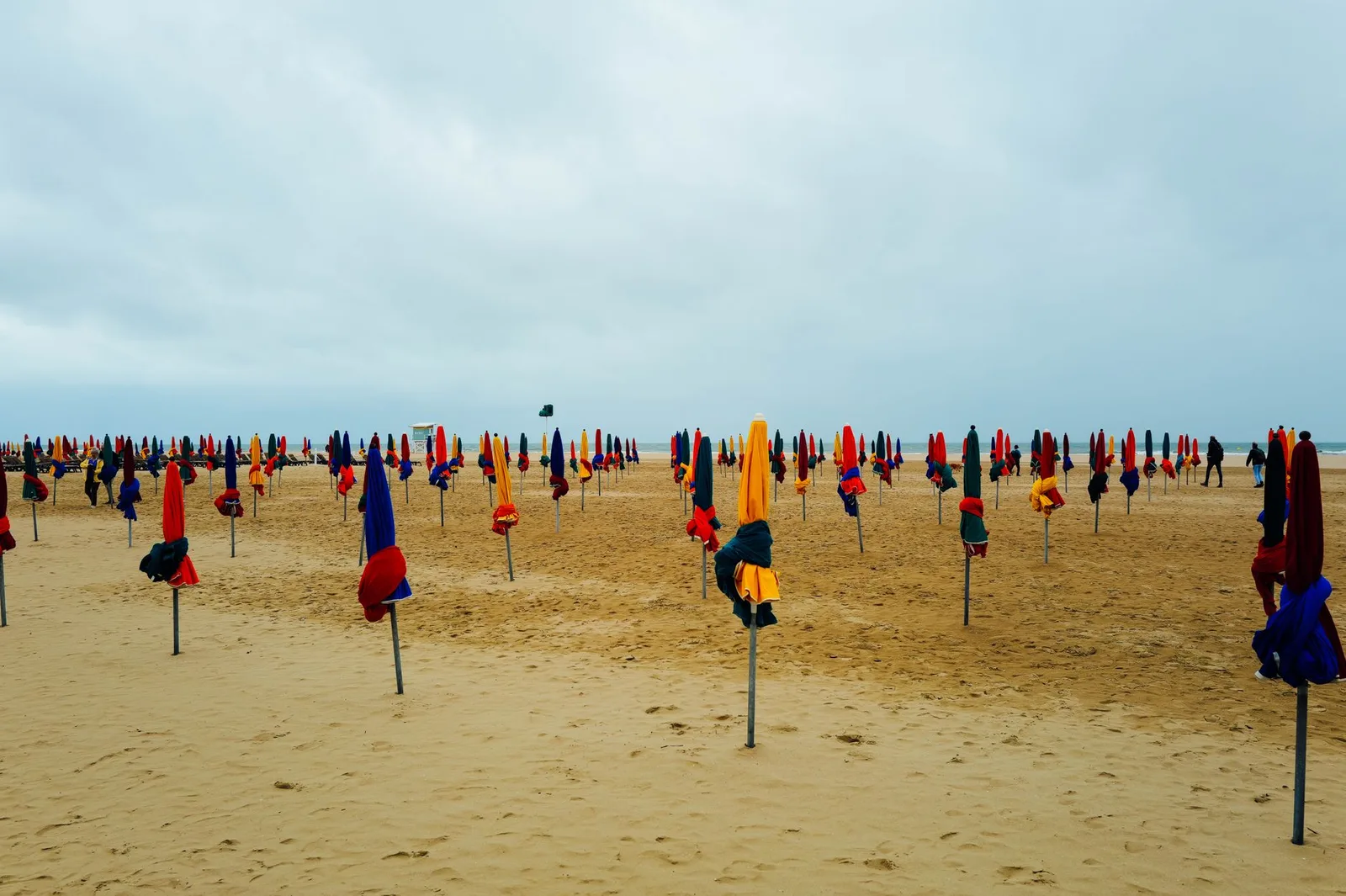 Reihen bunter Schirme stehen bereit am sandigen Strand von Deauville unter bewölktem Himmel.