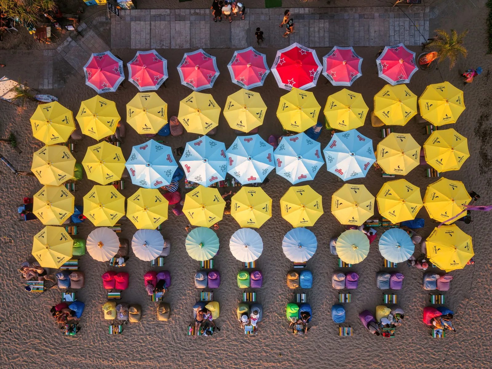 Aerial view of colorful beach umbrellas and seats on Bali's sandy shore during the day.