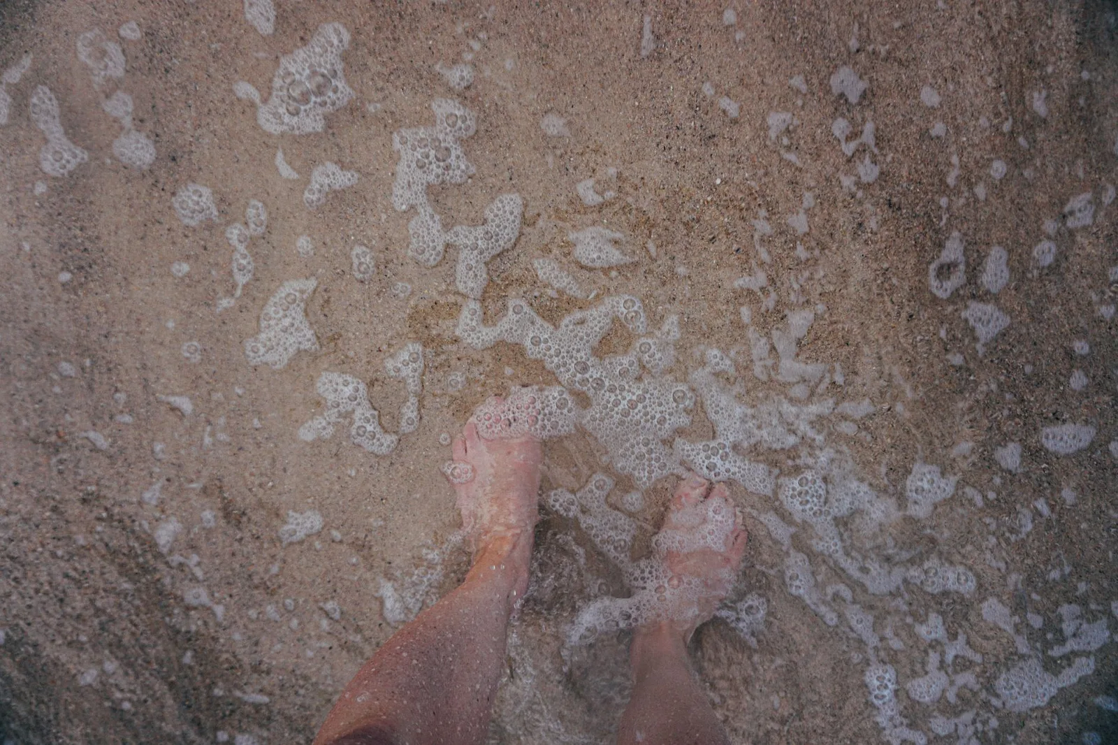 Top view of crop anonymous barefoot male tourist standing on sandy coast with transparent water